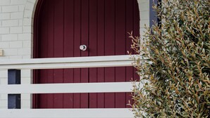 A red door with a round window, white railings, and a green bush in the foreground.