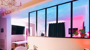A modern room with a round wooden table, white chairs, a chandelier, and a large window with curtains. - Dream Room | Jacuzzi, Sauna, Waterbed (Forbach)