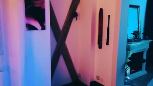 A room with a wooden floor, a large X-shaped black object on the wall, and a small table with a plant. - Dream Room | Jacuzzi, Sauna, Waterbed (Forbach)