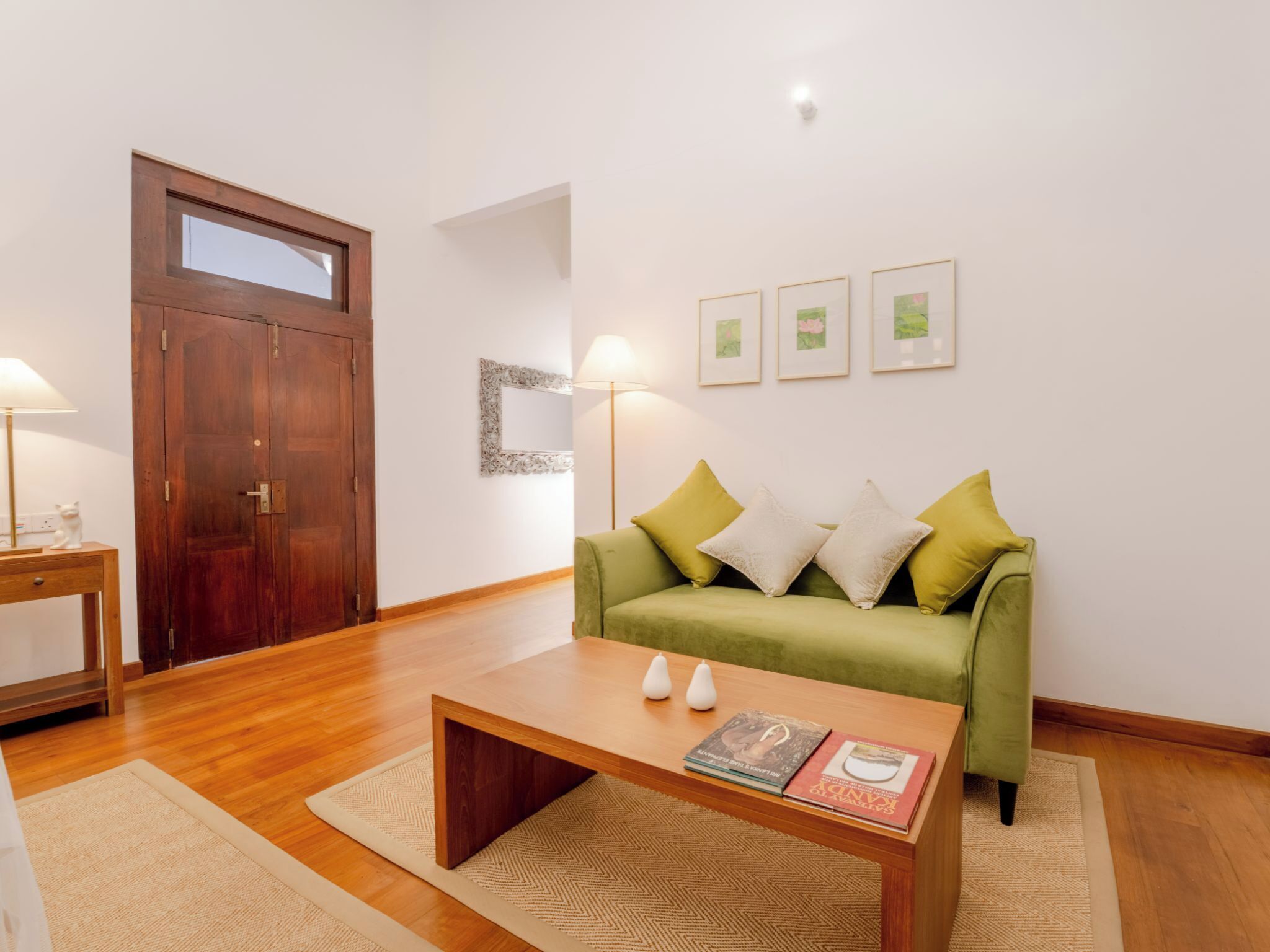 A modern living room with a wooden floor, a green sofa with cushions, a wooden coffee table with books, and a wooden door.