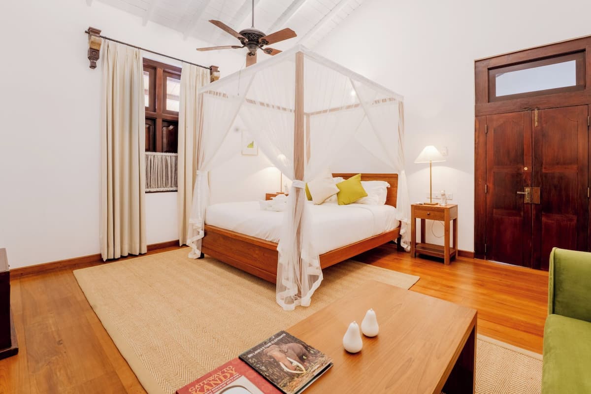 A bedroom with a four-poster bed, a ceiling fan, a wooden door, and a bedside table with a lamp.