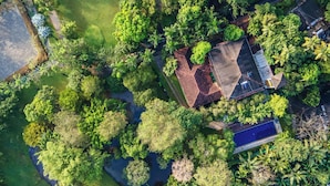 Vista aérea de una zona residencial con una casa de techo inclinado, rodeada de abundante vegetación y un pequeño estanque.