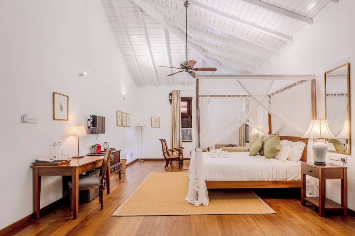 A spacious bedroom with a wooden floor, a bed with a canopy, a desk with a chair, a television, and a ceiling fan.