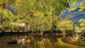 Un entorno tranquilo a la orilla del río, con árboles, un puente de madera y un edificio al fondo.