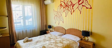 A hotel room with a large bed, two bedside tables, a chandelier, an air conditioning unit, and a decorative wall mural.