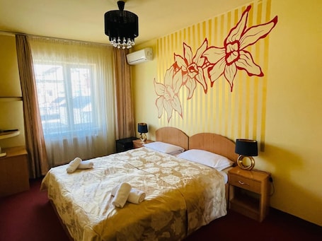 A hotel room with a large bed, two bedside tables, a chandelier, an air conditioning unit, and a decorative wall mural.