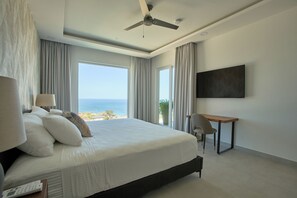 A bedroom with a bed, a desk, a chair, a TV, and a view of the ocean.