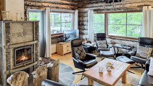 A log cabin interior with a stone fireplace, leather furniture, and large windows offering a forest view.