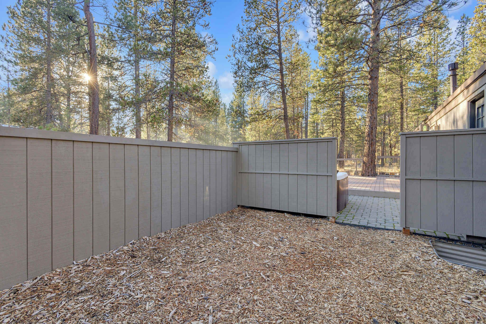 A fenced backyard with a wooden deck and a gravel area, surrounded by tall pine trees.