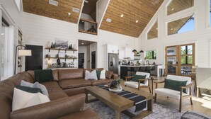 A spacious living area with a high ceiling, wooden beams, a brown leather sofa, and a kitchen in the background. - Luxury Forest Escape | NEW POOL, Loft, Swings Fire Pit &  gorgeous nature views (Broken Bow)