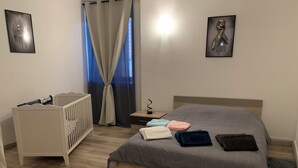 A bedroom with a wooden floor, a bed with a grey patterned bedspread, a white crib, a nightstand with a lamp, and two framed pictures on the walls.