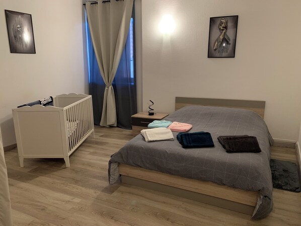 A bedroom with a wooden floor, a bed with a grey patterned bedspread, a white crib, a nightstand with a lamp, and two framed pictures on the walls.