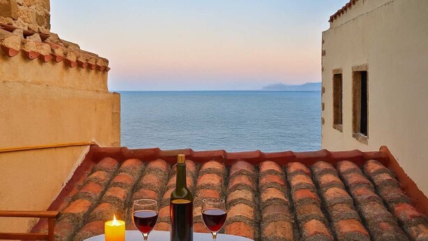 Une table avec deux verres de vin rouge et une bougie, sur une terrasse donnant sur la mer.