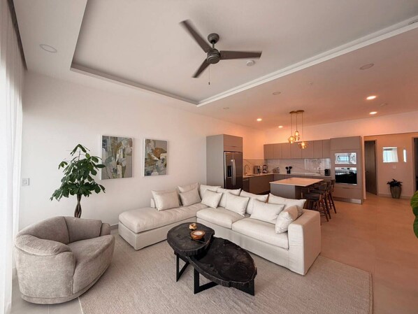 Un salon moderne avec un canapé, un fauteuil et une table basse, une cuisine avec un coin repas et un ventilateur de plafond.