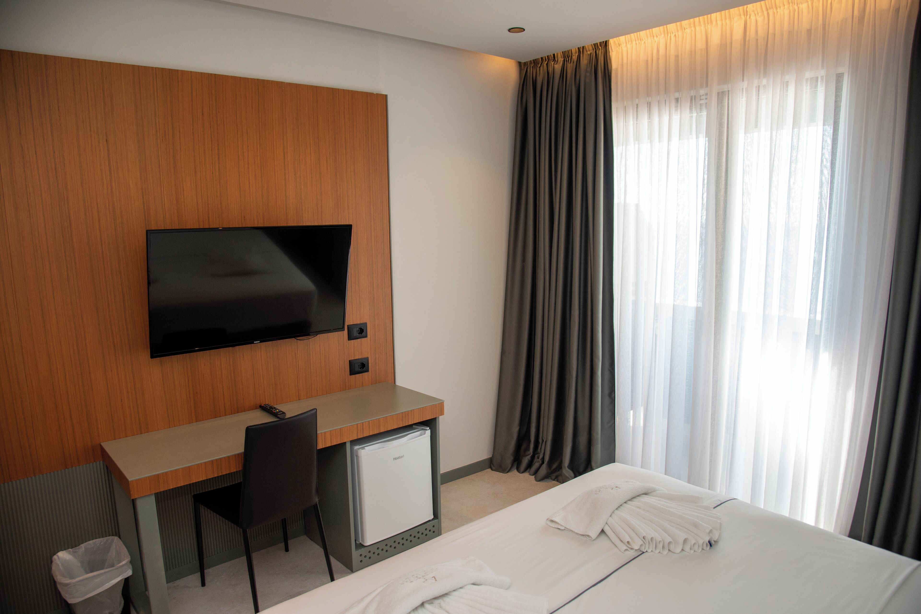 A hotel room with a bed, a desk with a television, a small refrigerator, and a window with curtains.