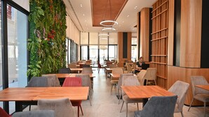 A modern restaurant with wooden tables and chairs, a green wall with plants, and a large window. - Adria Palace Hotel (Rrashbull)