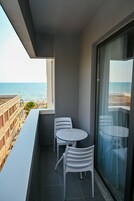 Un balcón con dos sillas blancas y una mesa, con vista a una playa y un edificio con balcones.