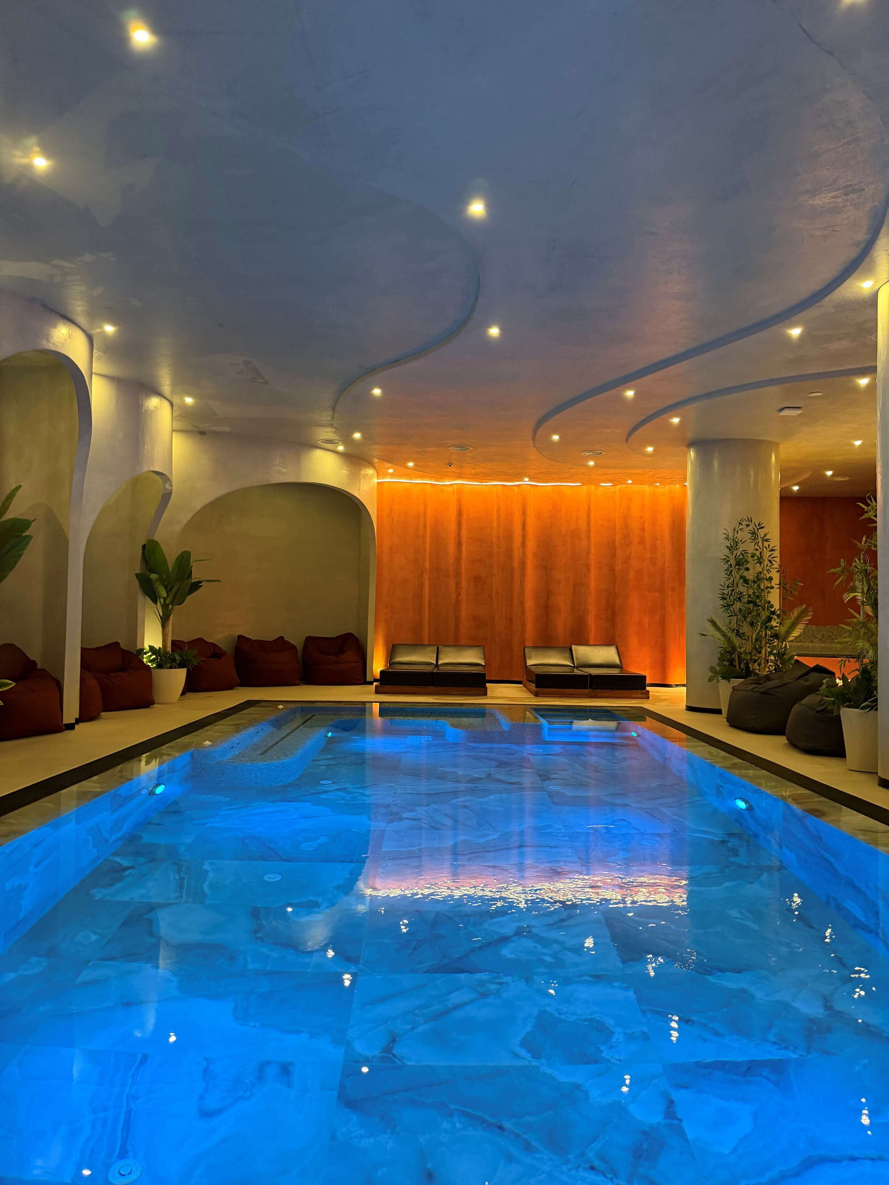A modern indoor pool with a blue-lit water surface, surrounded by seating areas with red cushions and potted plants.