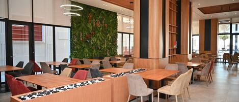 A modern restaurant with wooden tables and chairs, a green wall with plants, and large windows.