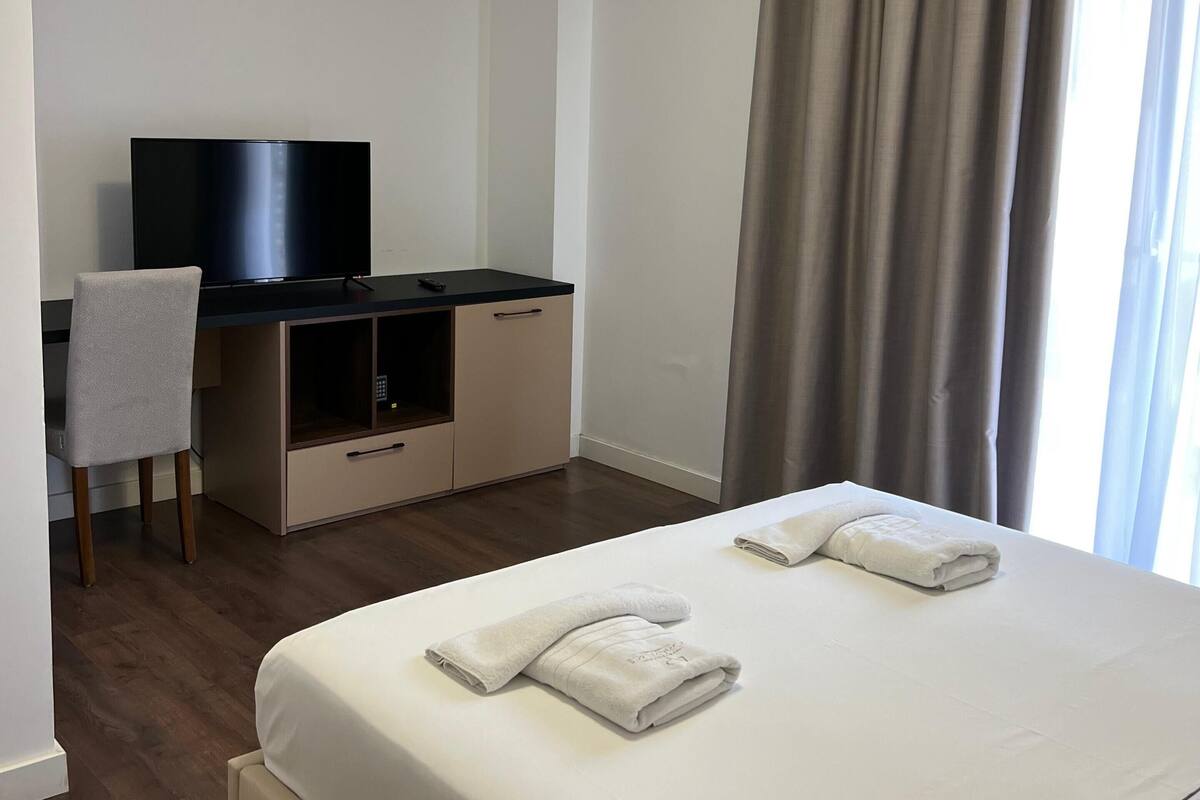 Ett hotellrum med en säng, ett skrivbord med en tv, en stol och ett fönster med gardiner.