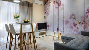 A modern living room with a grey sofa, a TV on a white stand, a small white table, and a floral wallpaper. - Casa Gardenia -modern apartment next to the beach and near old town (Chania)