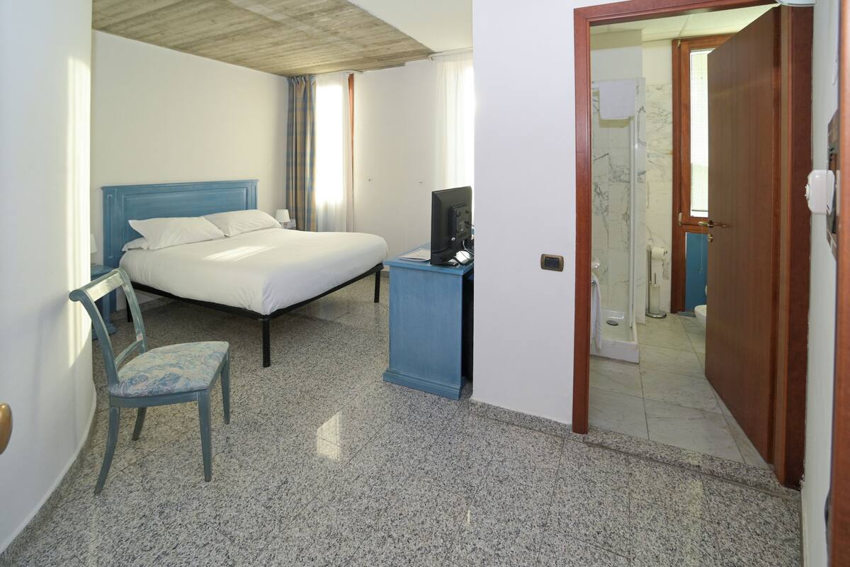 Un dormitorio con cama, una silla, televisor sobre un soporte y un baño visible a través de una puerta abierta.
