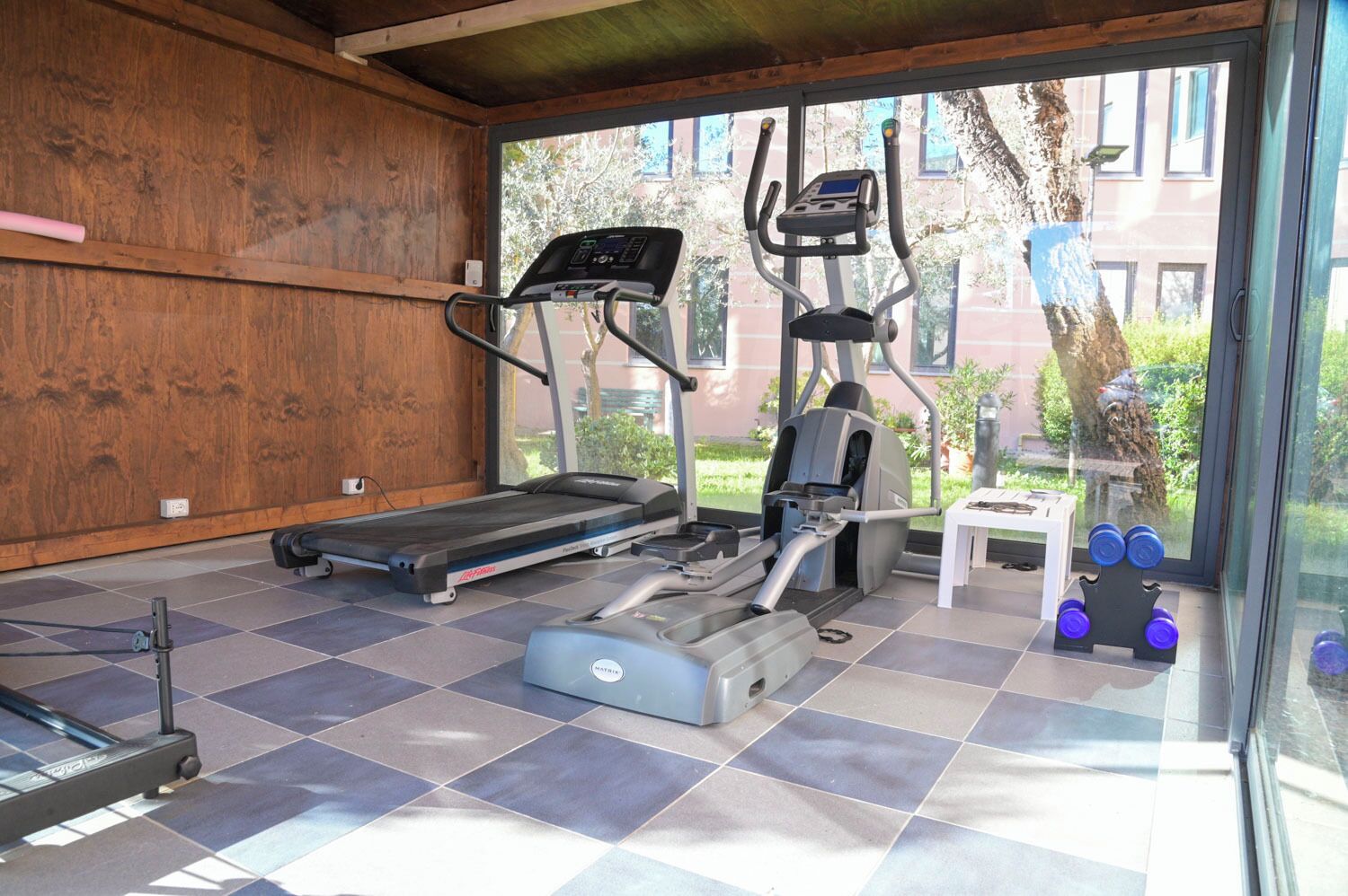 A gym with a treadmill, elliptical, and weights, with a view of a building and trees outside.