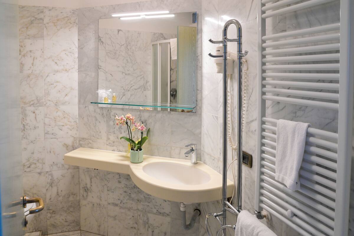 Un baño con pared de mármol, repisas de vidrio con artículos de aseo, un lavamanos, un espejo, un perchero para toallas y una ducha con cabezal de ducha manual.