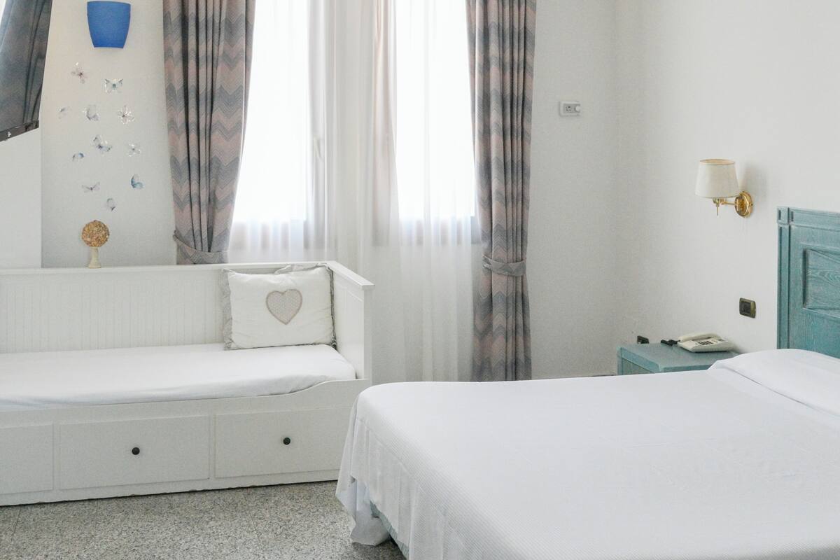 Un dormitorio con una cama blanca, una mesita de noche con lámpara, un tocador pequeño y una ventana con cortinas.