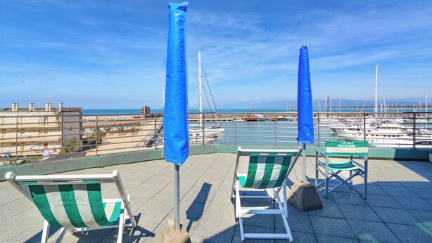 Une terrasse sur le toit, dotée de deux chaises longues à rayures vertes et de parasols bleus, qui offre une vue sur une marina avec des bateaux et un ciel bleu dégagé.