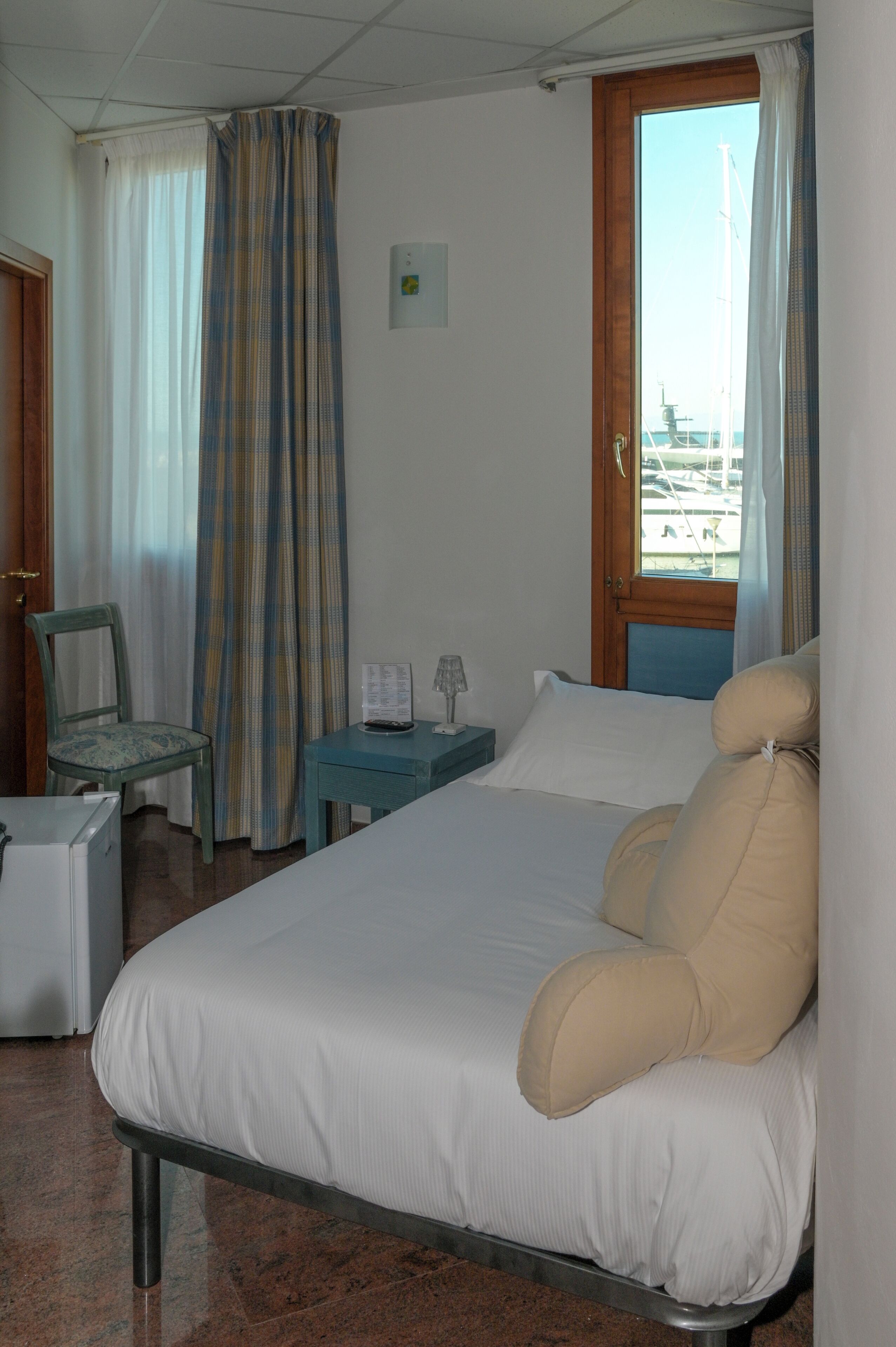 Habitación de hotel con cama, una silla, una mesita de noche con lámpara y ventana con cortinas.
