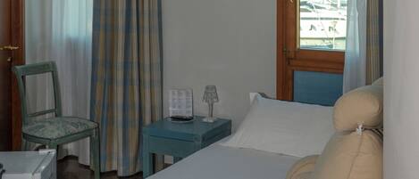 Habitación de hotel con cama, una silla, una mesita de noche con lámpara y ventana con cortinas.