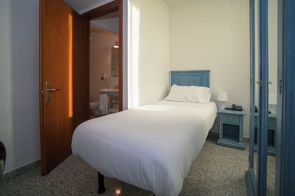 Una habitación de hotel con puerta de madera, cama con ropa de cama blanca, mesita de noche pequeña con lámpara, y baño visible a través de una puerta abierta.