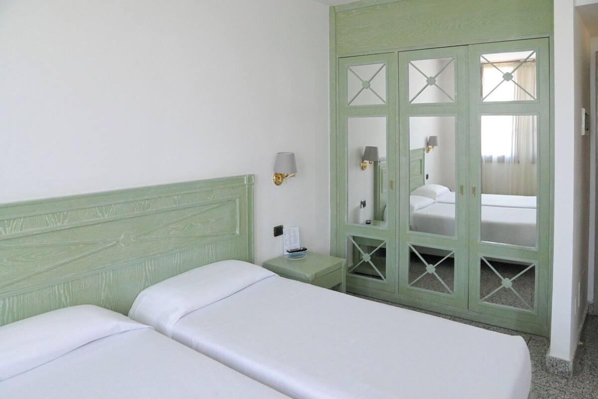 Una habitación con dos camas, un armario verde con espejo, una lámpara de noche y una pequeña mesa.