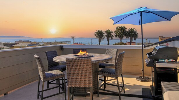 A rooftop terrace with a round table, chairs, a fire pit, a grill, and a view of the sunset over the ocean. - Steps to Surf & Sand | Beachfront Oasis + Pier Walk (Pismo Beach)