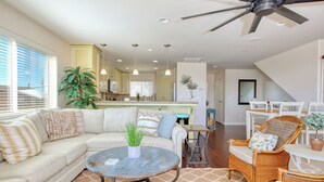 A spacious living area with a sofa, coffee table, and wicker chairs. A ceiling fan is visible, and there is a kitchen in the background. - Steps to Surf & Sand | Beachfront Oasis + Pier Walk (Pismo Beach)
