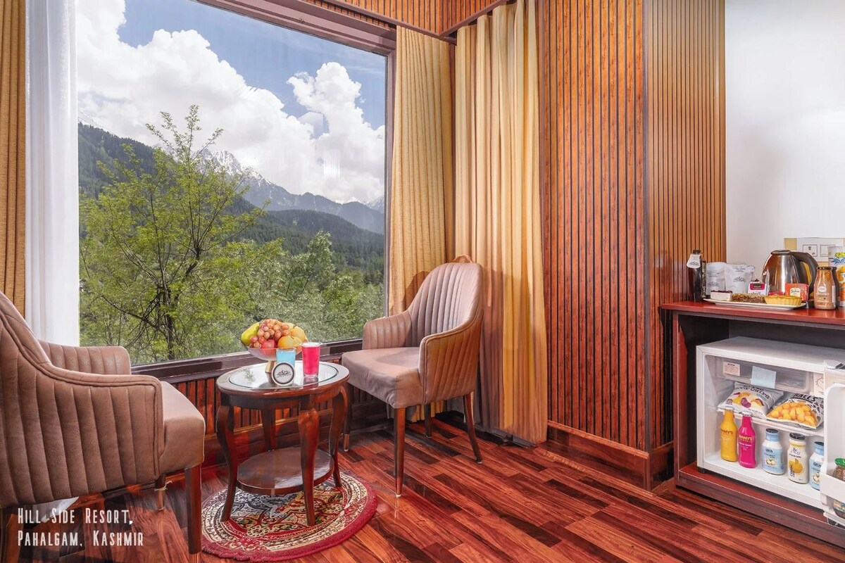 A room with a large window offering a mountain view, two chairs, a small table with a fruit bowl, and a minibar with beverages.