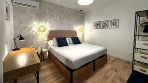 Un dormitorio con cama, mesita de noche, lámpara, aire acondicionado instalado en la pared, luz de techo y cuadros enmarcados.