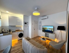Un salon moderne comprenant un canapé, des fauteuils, un téléviseur et un coin cuisine équipé d’appareils électroménagers.