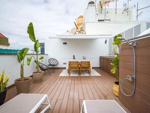 Una terraza en la azotea con cubierta de madera, dos mesas de madera, sillas, plantas en macetas, un rincón de descanso y una zona de ducha.