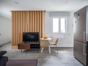 Un espace de vie moderne comprenant un téléviseur à écran plat, une table à manger avec des chaises, un réfrigérateur et une fenêtre avec des stores.