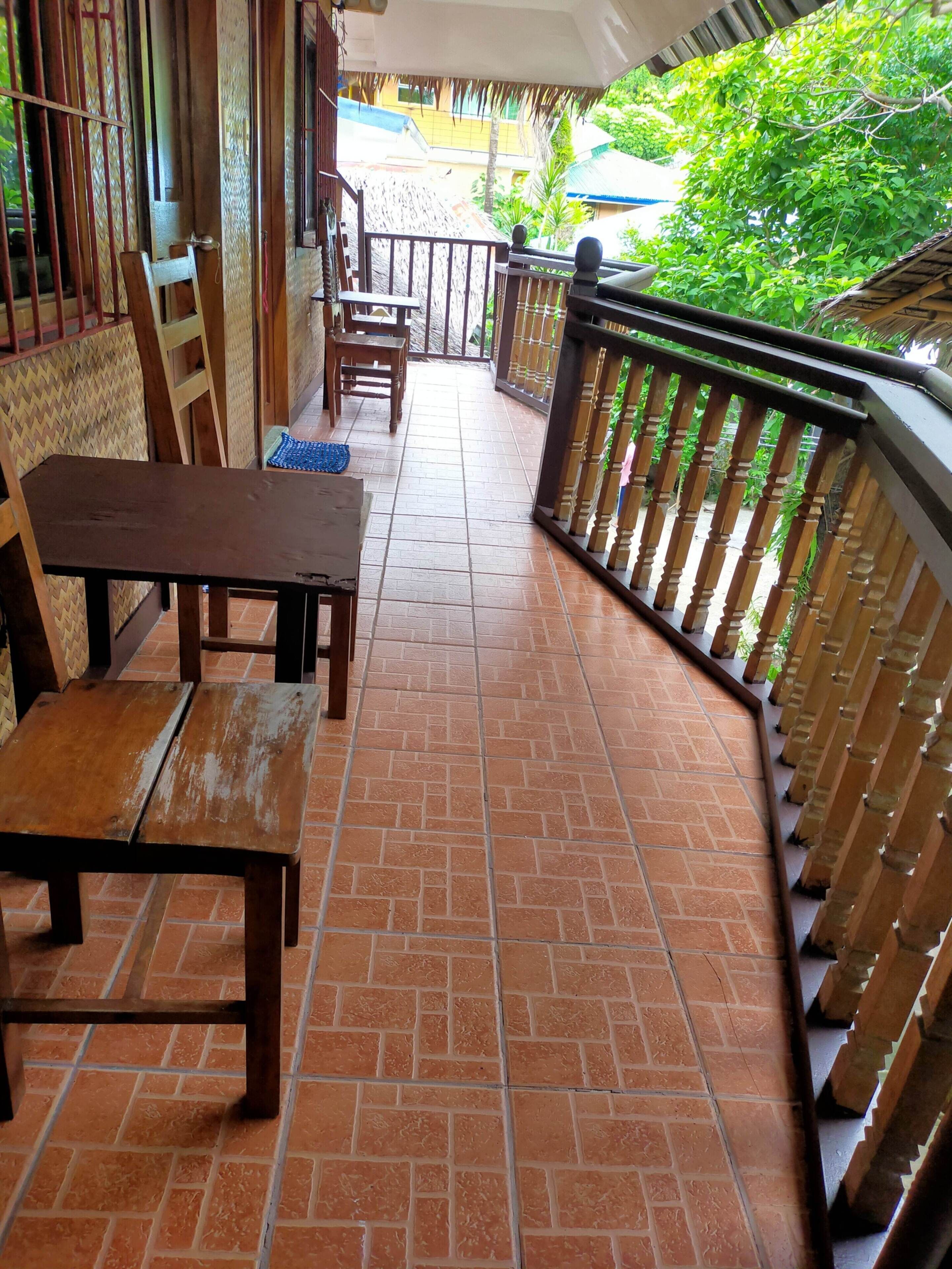 A covered outdoor area with tiled flooring, wooden furniture, and a railing overlooking greenery.