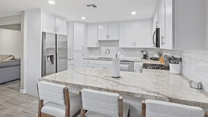 A modern kitchen with white cabinets, a granite countertop, and a stainless steel refrigerator. There are three bar stools in front of the counter. - Solace | Pool, Hot Tub, Cabana, Fire Pit, Outdoor Living, Sleeps 22 | Old Town Scottsdale (Scottsdale)