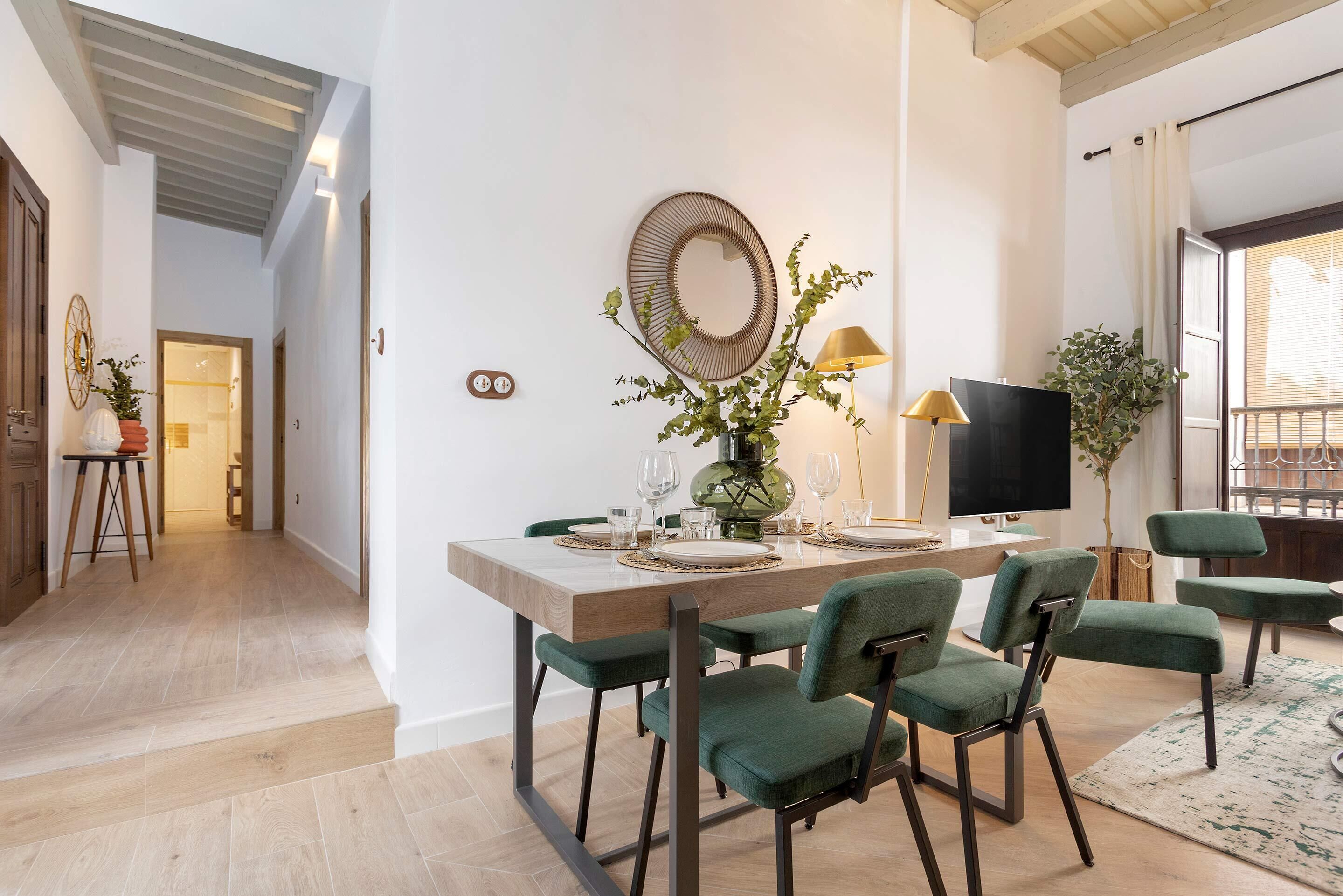 A dining area with a wooden table, green upholstered chairs, a decorative mirror, and a television on a stand.
