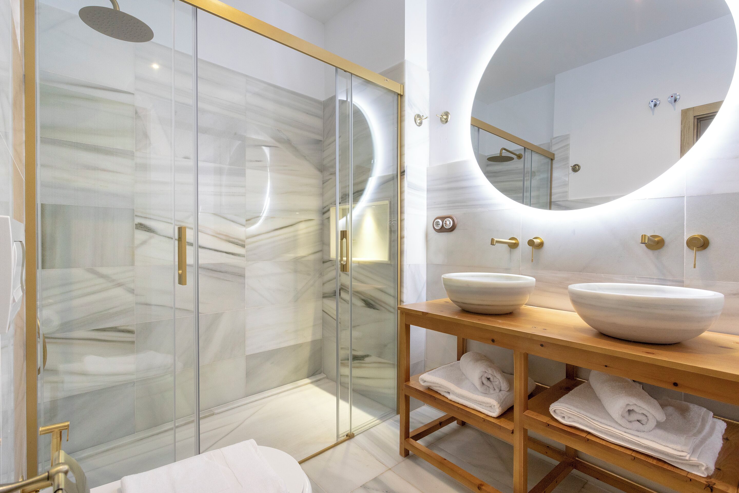 A modern bathroom with a glass-enclosed shower, marble walls, a wooden vanity with two bowls, and a round mirror.