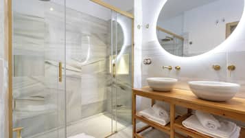 A modern bathroom with a glass-enclosed shower, marble walls, a wooden vanity with two bowls, and a round mirror.
