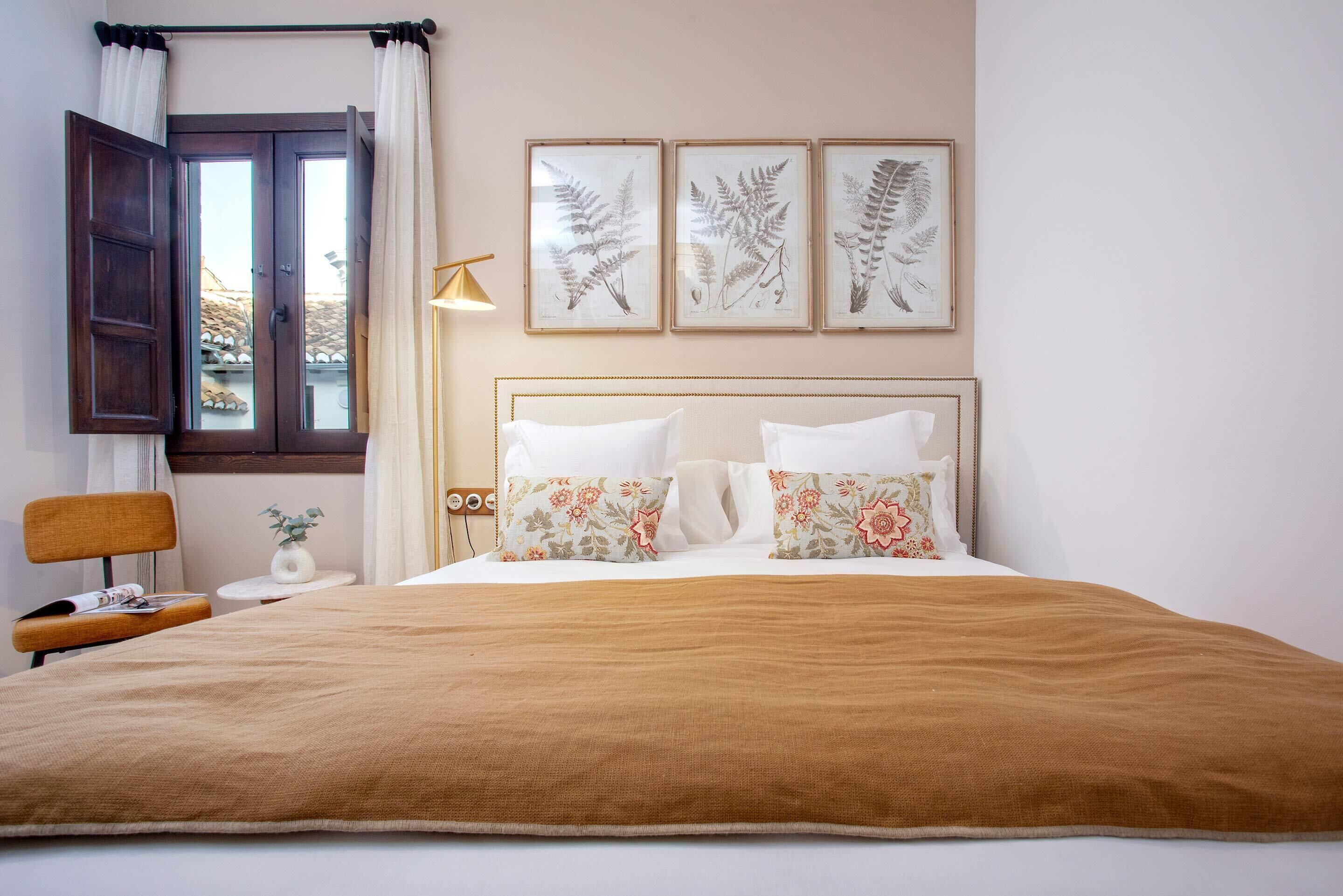 A bedroom with a bed, a window with curtains, a bedside table with a lamp, and framed botanical prints on the wall.