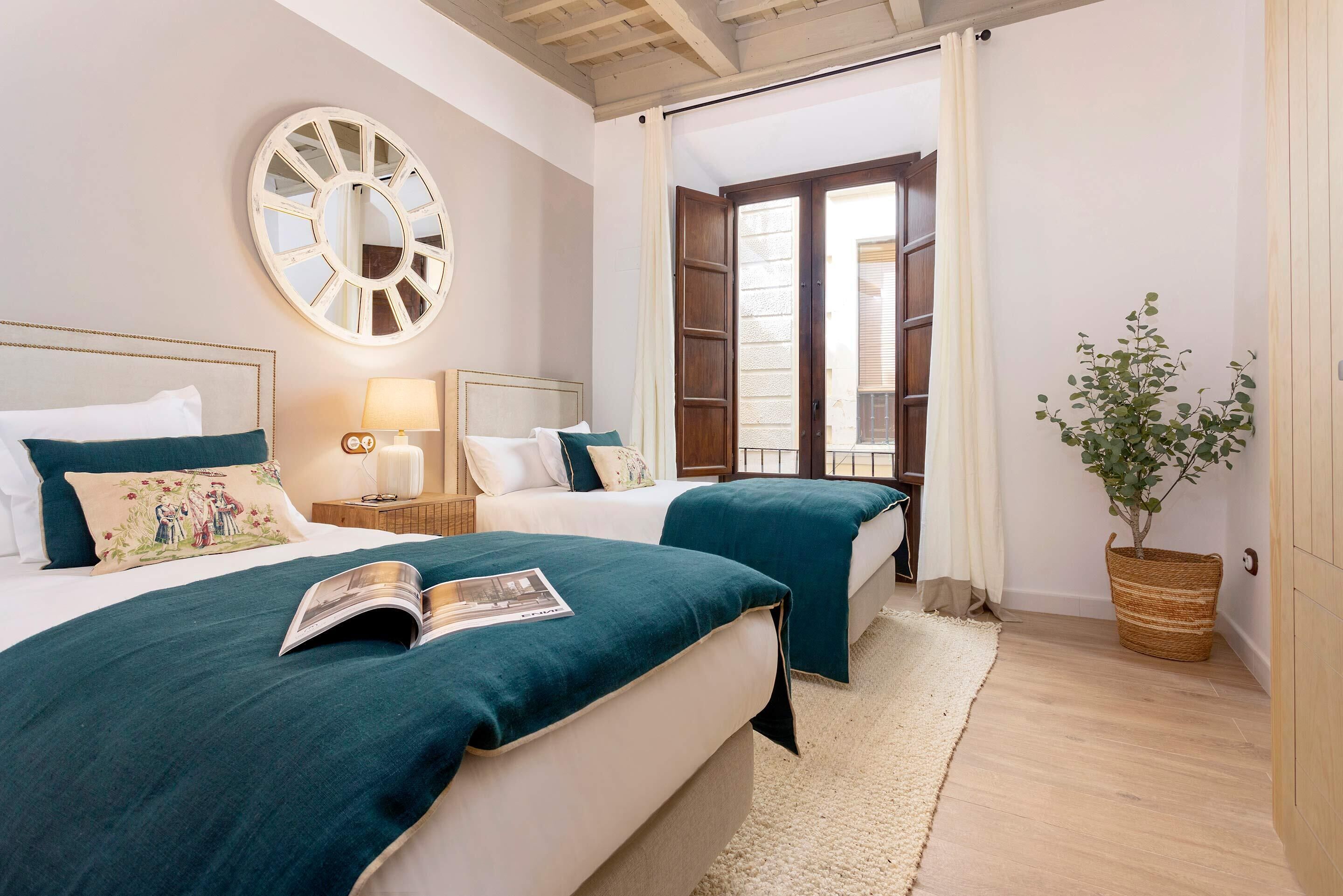A bedroom with two beds, a large mirror, a potted plant, and a view through an open door.