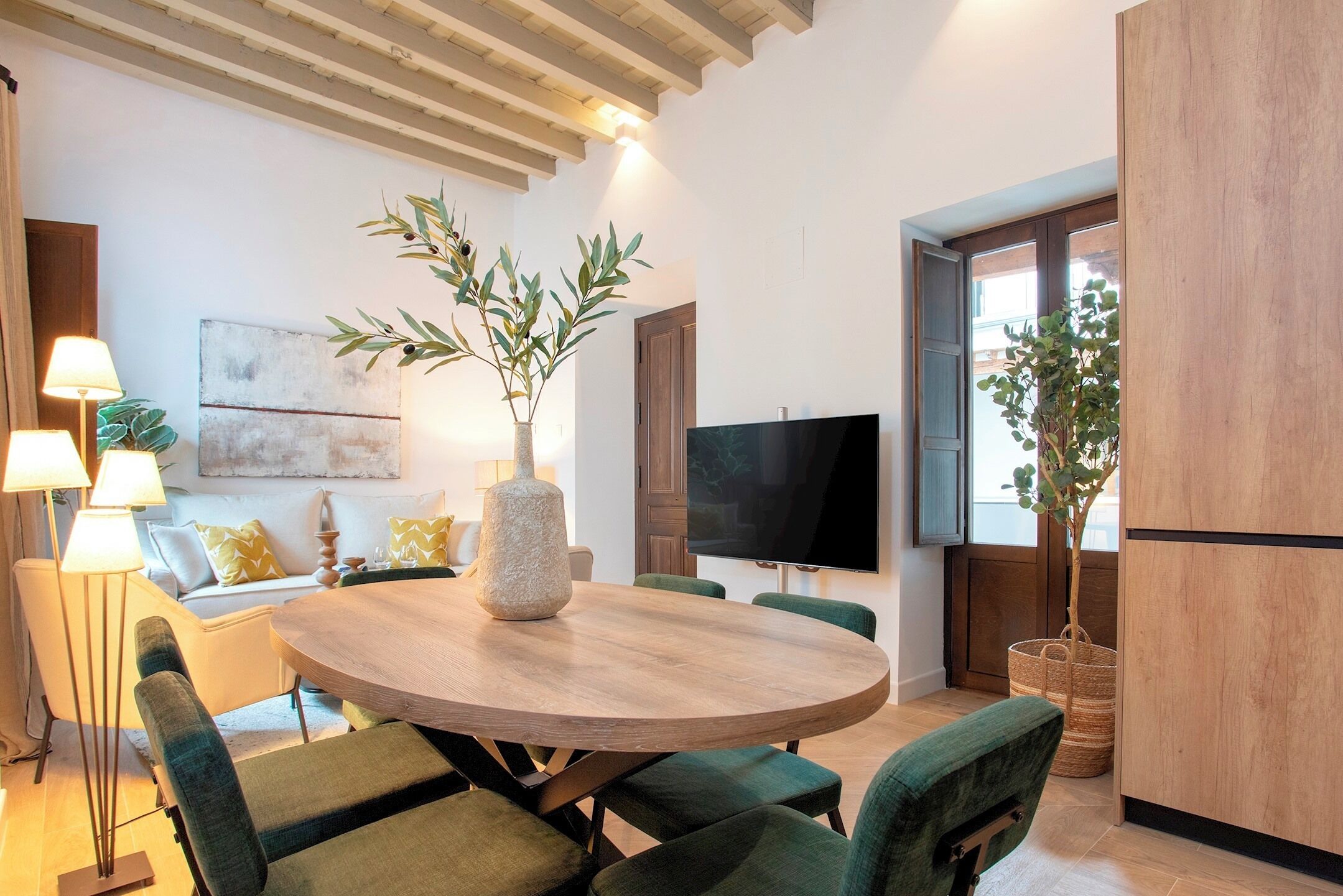 Un salon moderne avec une table ronde en bois, des fauteuils à revêtement vert, un grand téléviseur à écran plat et une plante en pot.