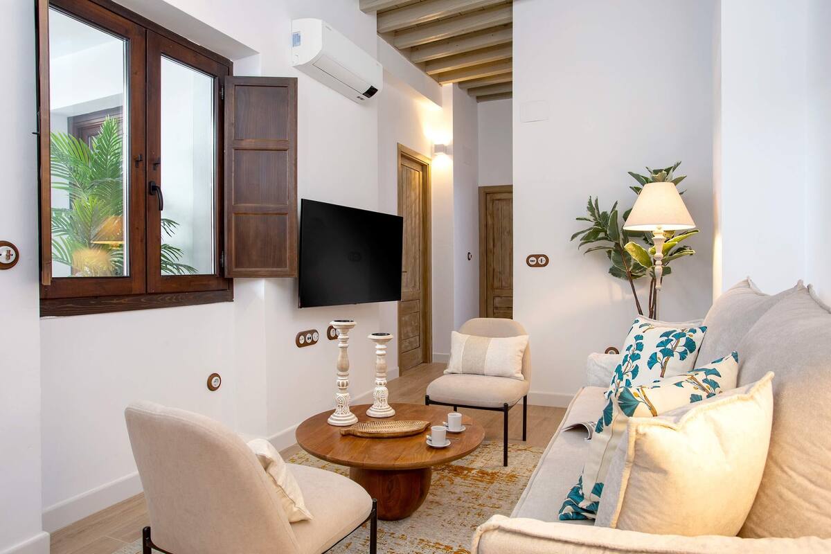 Ein Wohnzimmer mit einem runden Holztisch, beigen Sesseln, einem Sofa mit floralen Kissen, einem an der Wand befestigten Fernseher und einer Lampe.